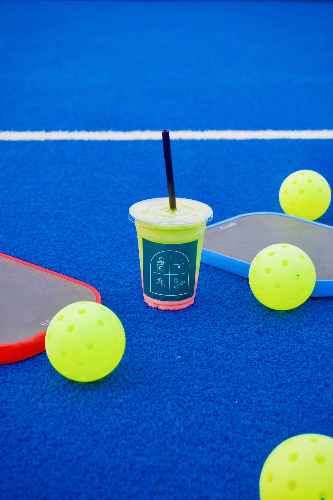 Alkemy iced drink on a blue pickleball court with paddles and balls